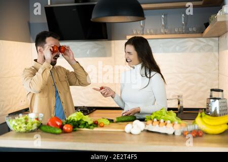 Cute couple are smiling and having fun in kitchen. Happy vegan people are cooking salad using only nutrition and healthy food. Stock Photo