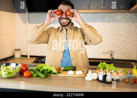 White man with beard is smiling and having fun in kitchen. Happy vegan man is cooking salad using only nutrition and healthy food. Stock Photo