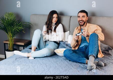 Sweet couple has quarantine free time play video games. She doesn't like and disappointed felling bored while he is having fun playing with pop corn b Stock Photo