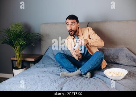 Young attractive man playing console game in front of camera, beating his opponent online. White man on sofa with pop corn. E-sport competition concep Stock Photo