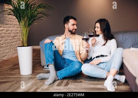 Young couple is sitting on the floor with red wine in bedroom and celebrating their new apartment. Stock Photo