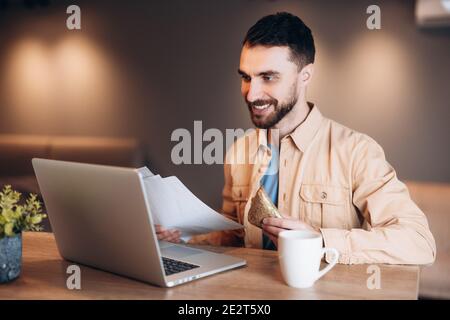 Freelancer man eating sandwich, drinking coffee and meeting with boss by videocall. Man using his laptop and smiling. Working from home. Casual style. Stock Photo