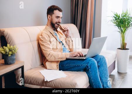 Man watching funny videos on laptop. Good looking young man in casual clothes eats lunch while working from home. Quarantine concept. Living room on Stock Photo