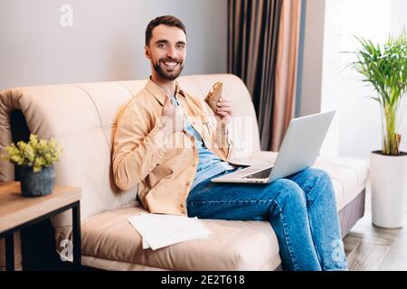 I really enjoy this leisure. Cheerful man in orange shirt and blue jeans makes thumbs up gesture and holds sandwich in another. Modern laptop lying on Stock Photo