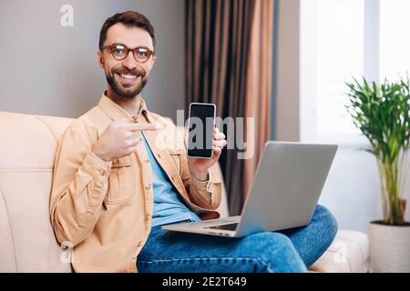 Male freelancer is happy to work from home. Good looking smart man with bright smile holds smartphone in one hand and points at it with another Stock Photo