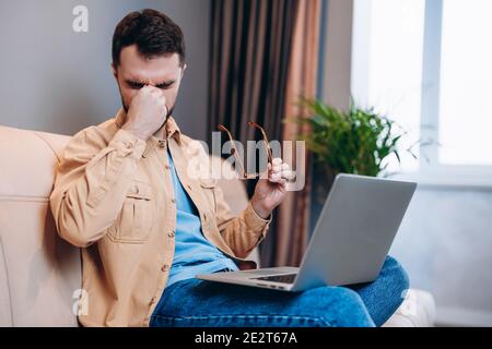 Exhausted freelancer working from home. Tired good looking man with beard holds his head with painful look on his face. Remote work concept. Hard to Stock Photo