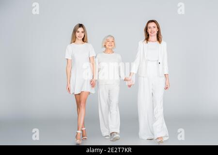 full length of three generation of happy women smiling while holding hands and walking on grey Stock Photo
