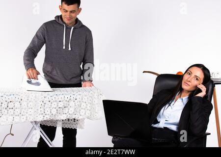A young business woman decides business issues by phone while sitting at a computer. The man ironing clothes. Stock Photo
