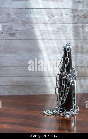 beer bottle with chains illuminated by a ray of sunshine with a wooden base as a measure of closure for hotels and bars for covid19 and alcoholism Stock Photo