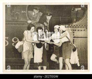 WW2 era press photo of happy young female walkers reading a map at train station platform, beside a steam engine, train drivers look on, London, U.K., 1940's Stock Photo