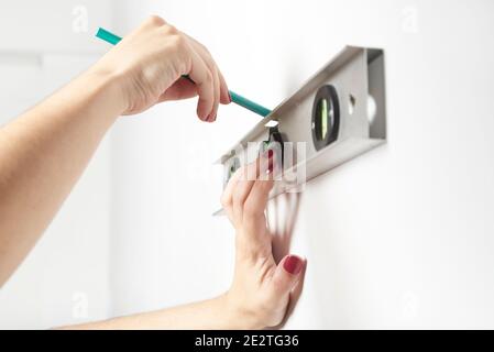 Young woman with manicured hands making a mark on a white wall using a pencil and a bubble level. Concepts of renovating and decorating. Stock Photo