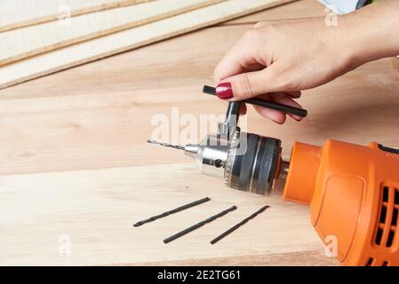 Manicured hand of a young woman changing the drill bit of an electric drill on a wooden table. Female using construction tools. Image with copy space. Stock Photo