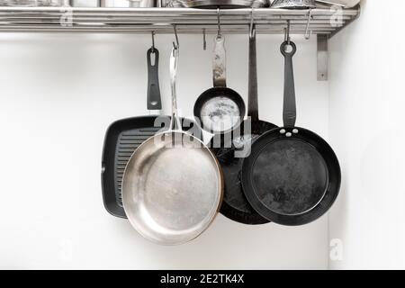 Various pans in different sizes and forms for cooking and frying hanging on metal hooks from shelf in kitchen with white wall in background