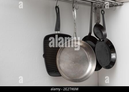 Various pans in different sizes and forms for cooking and frying hanging on metal hooks from shelf in kitchen with white wall in background