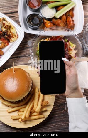 Female make a online order on a smartphone. Food container on a wooden table. Fast food delivery at home. Eating business lunch at home. Work from home. Remote work Stock Photo
