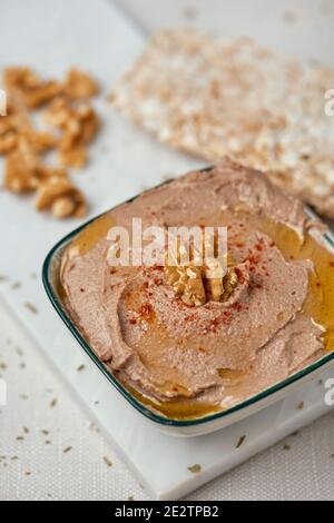 a white ceramic bowl with an appetizing hummus, made with chickpeas, walnuts and rosemary, next to some puffed rice cakes on a white table Stock Photo