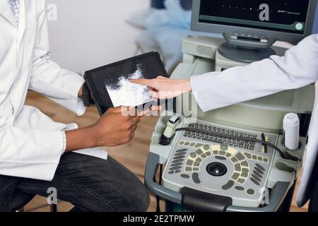 Cropped top view of black male doctor hands, holding ipad pc with ultrasound scan, while female hand of patient or doctor colleague pointing on tablet Stock Photo