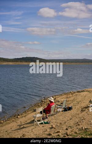 Caucasian senior woman sitting on a chair with a straw hat on a dam lake reservoir in Alentejo, Portugal Stock Photo