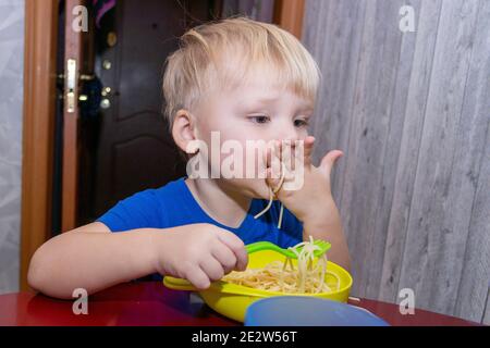 a small child eats noodles in the kitchen, helping himself with his hands Stock Photo