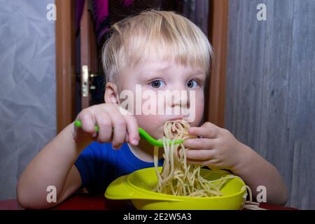 a small child eats noodles in the kitchen, helping himself with his hands Stock Photo