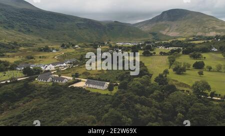Scottish village cottages, houses at road aerial. Whiskey distillery at mountain green valley. Nobody nature landscape at summer cloudy day. High mounts of Arran Island, Scotland. Cinematic shot Stock Photo