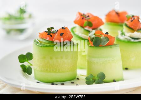 Holiday vegetable appetizers. Cucumbers rolls with soft cheese, pieces of salted salmon, microgreens and black sesame served on a white plate. Selecti Stock Photo