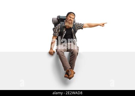 Bearded man with a backpack and hiking equipment sitting on a panel and pointing to the side isolated on white background Stock Photo