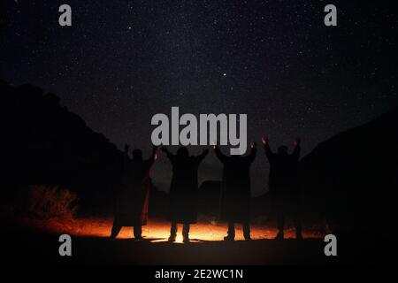 Four persons wearing long warm coats standing in night desert, hands raised to sky with stars, light on the ground, view from behind only silhouettes Stock Photo