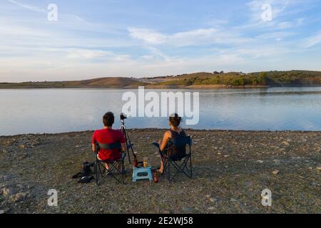 Caucasian couple sit on chairs on a lake drinking beer and seeing the landscape in Alentejo, Portugal Stock Photo