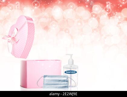 Valentine's Day concept during Covid-19 pandemic with opened heart-shaped pink gift box, face mask, hand sanitizer and copy space on pink bokeh backgr Stock Photo