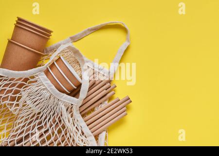 Drinking brown tubes straws made of paper and cornstarch, mesh market bag and empty paper coffee cups on a trendy grey and yellow background. Zero was Stock Photo