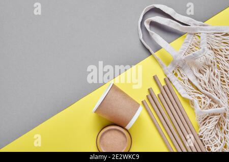 Drinking brown tubes straws made of paper and cornstarch, mesh market bag and empty paper coffee cups on a trendy grey and yellow background. Zero was Stock Photo