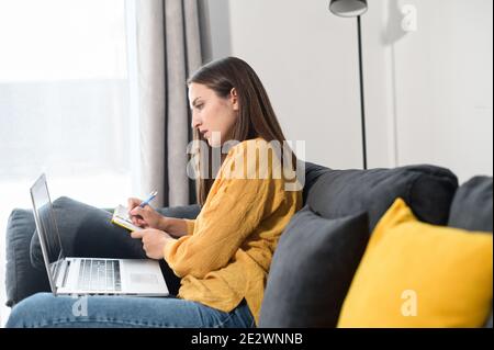 Focused young woman works with a laptop sitting on the couch at home, female student watching a webinar, video courses and taking a notes in a notepad Stock Photo