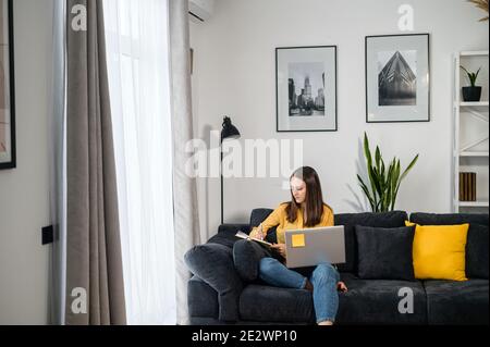 Focused young woman works with a laptop sitting on the couch at home, female student watching a webinar, video courses and taking a notes in a notepad Stock Photo