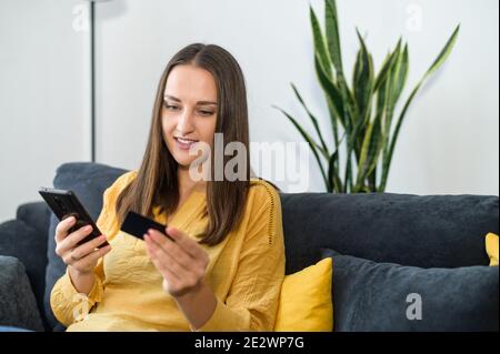 A smiling woman sits on the sofa with a modern smartphone and credit card in the hands shopping and paying online. A young freelancer receives Stock Photo