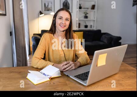 A young cheerful woman using headset for online talking sitting at the desk at home office, pleasant female operator looks at camera. Remote work Stock Photo