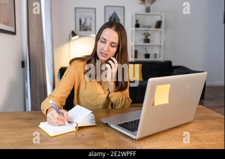 A young female freelancer talking on the phone and using a laptop working remotely at home office. A woman in yellow casual shirt taking notes Stock Photo