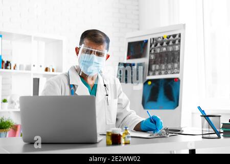 Middle-aged male doctor in face shield and mask sitting at the table at his office Stock Photo