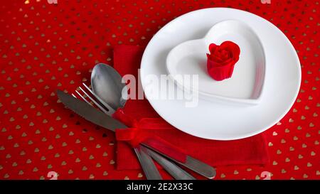 Romantic decoration of holiday table. White plates in the shape of a heart with a red rose. Red background Stock Photo
