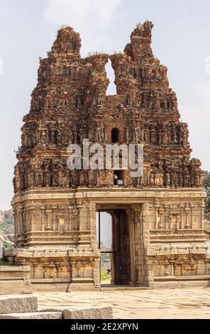 Hampi, Karnataka, India - November 5, 2013: Vijaya Vitthala Temple. Damaged red stone Gopuram on top of yellow stone entrance gate. Some statues barel Stock Photo