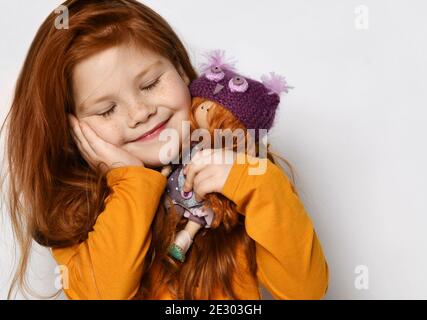 Cute five-year-old red-haired kid girl with closed eyes in orange sweatshirt holds small redhair doll in hands at face Stock Photo