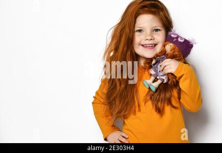 Frolic smiling five-year-old red-haired kid girl in orange sweatshirt holds small redhair doll in hands on her shoulder Stock Photo