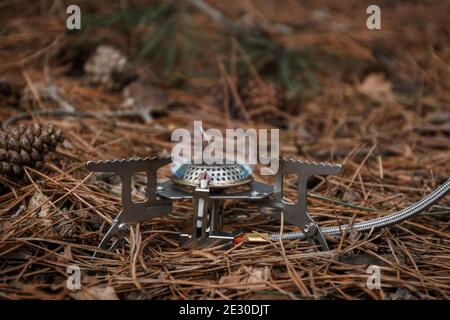 Tourist gas burner for heating food. Open fire close-up. Metal portable small burner in the forest on coniferous litter.The concept of hiking, tourism Stock Photo