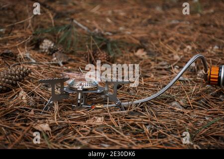 Tourist gas burner for heating food. Open fire close-up. Metal portable small burner in the forest on coniferous litter.The concept of hiking, tourism Stock Photo