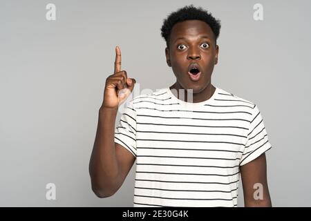 Stupefied afro young man makes big eyes opens mouth widely, raise finger up, having great idea.  Stock Photo