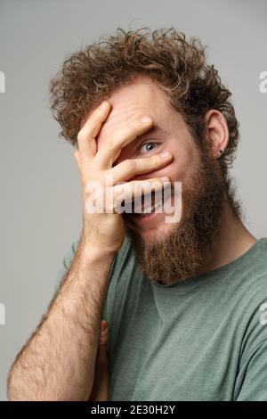 Shy handsome young man covered his face with hand pry with his eye, curly hair in olive t-shirt isolated on white background. Portrait of smiling Stock Photo