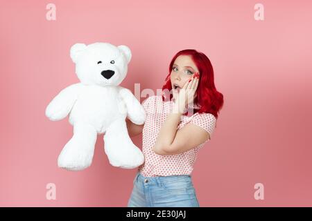 close-up a surprised, shocked young woman with an open mouth and red hair holds a large white teddy bear isolated on a pink background Stock Photo