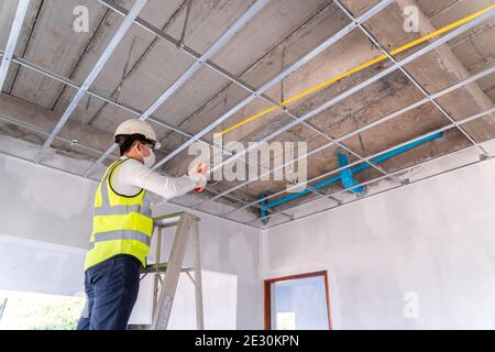 Electrician wearing a mask installing laying electrical cables on the ceiling with pliers inside the house under construction. Stock Photo