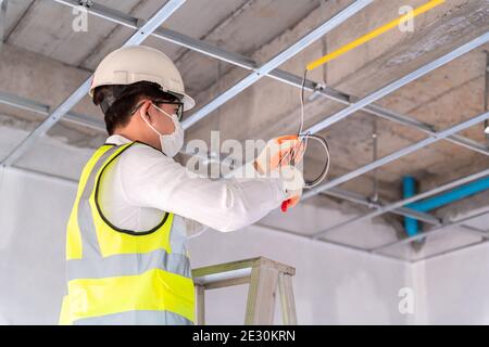 Electrician wearing a mask installing laying electrical cables on the ceiling with pliers inside the house under construction. Stock Photo