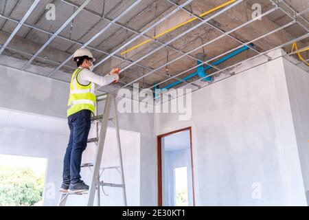 Asian electrician wearing a mask installing laying electrical cables on the ceiling with pliers inside the house under construction. Stock Photo